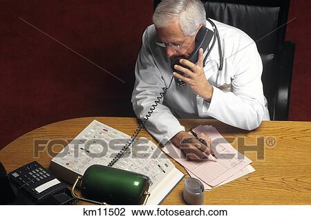Stock Image - 1990S Senior Doctor At Desk Talking On Telephone And Writing On Chart . Fotosearch