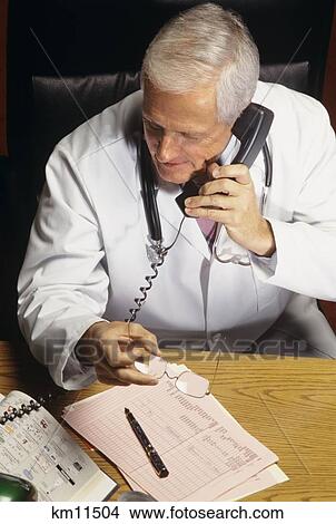 Picture - 1990S Senior Doctor Sitting At Desk Holding Glasses Talking On Telephone . Fotosearch