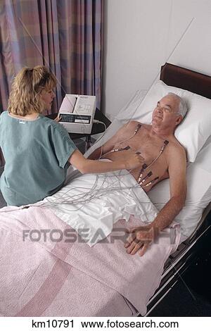 Stock Image - 1990S Technician Performs Ekg On Male Patient In Hospital Bed . Fotosearch