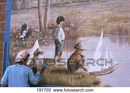 Stock Image - 1990S Woman Painting A Nostalgic Wall Mural Of Boys Sailing Toy Boat In Pond. Fotosearch