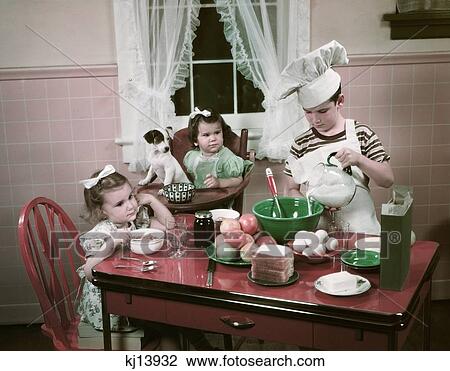 Stock Image - 1940S 1950S Children Two Girls At Kitchen Table With Puppy And Boy Cooking Wearing Chefs Toque. Fotosearch