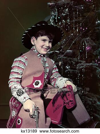 1940S 1950S Smiling Boy Dressed In Cowbyo Hat Costume Holding Toy Gun Sitting Wooden Horse By Christmas Tree View Large Photo Image Stock Photo - 1940S 1950S Smiling Boy Dressed In Cowbyo Hat Costume Holding Toy Gun Sitting Wooden Horse By Christmas Tree . Fotosearch