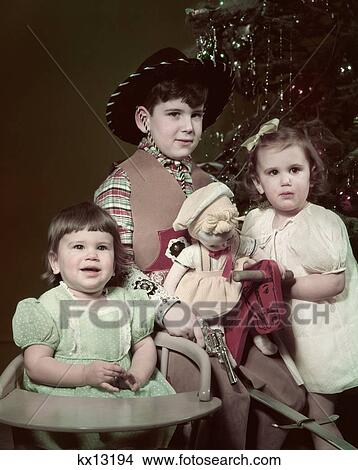 1940S 1950S Three Children By Christmas Tree Boy Cowboy Hat Costume Girl With Doll Toddler In Chair View Large Photo Image Picture - 1940S 1950S Three Children By Christmas Tree Boy Cowboy Hat Costume Girl With Doll Toddler In Chair . Fotosearch