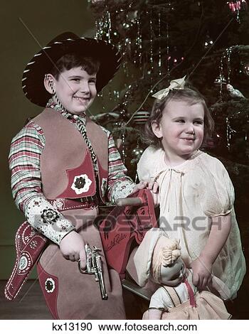 1950S Smiling Boy Dressed In Cowboy Hat Outfit Holding Cap Pistol Little Girl Holding A Doll By Christmas Tree View Large Photo Image Stock Image - 1950S Smiling Boy Dressed In Cowboy Hat Outfit Holding Cap Pistol Little Girl Holding A Doll By Christmas Tree . Fotosearch