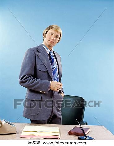 1960S 1970S Serious Blond Businessman Wearing Knit Suit Standing By Desk Looking At Camera View Large Photo Image Stock Photograph - 1960S 1970S Serious Blond Businessman Wearing Knit Suit Standing By Desk Looking At Camera. Fotosearch