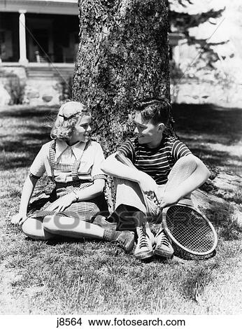 1930S 1940S Boy And Blond Girl Sitting Outside By Tree Trunk Talking Holding Tennis Racket View Large Photo Image Picture - 1930S 1940S Boy And Blond Girl Sitting Outside By Tree Trunk Talking Holding Tennis Racket . Fotosearch