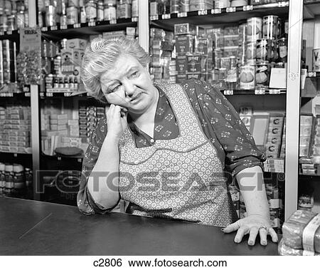 1930S 1940S Sad Senior Woman Clerk Grocery Store Wearing Apron Canned Goods On Shelves Leaning Counter View Large Photo Image Stock Photograph - 1930S 1940S Sad Senior Woman Clerk Grocery Store Wearing Apron Canned Goods On Shelves Leaning Counter. Fotosearch