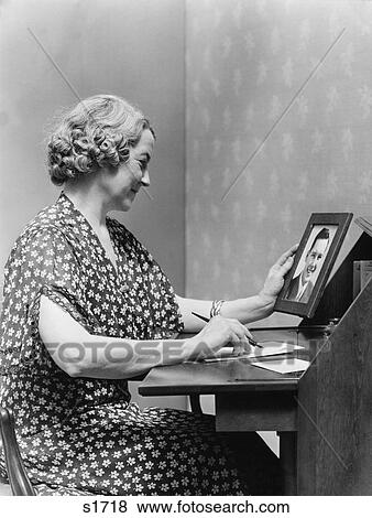 1930S 1940S Senior Woman Sitting At Desk Writing Letter As She Holds Picture Portrait Of Young Man Son View Large Photo Image Stock Photo - 1930S 1940S Senior Woman Sitting At Desk Writing Letter As She Holds Picture Portrait Of Young Man Son . Fotosearch