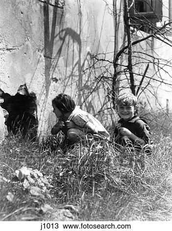 1930S 2 Kids Beside Old Building Girl Peeking Into Hole In Wall Boy Sitting In Grass Smiling View Large Photo Image Stock Image - 1930S 2 Kids Beside Old Building Girl Peeking Into Hole In Wall Boy Sitting In Grass Smiling . Fotosearch