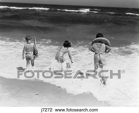 Stock Image - 1930S Three Children Two Boys One Girl Back View Stepping Into Ocean Surf Wave At Waters Edge Seashore Summer Fun. Fotosearch
