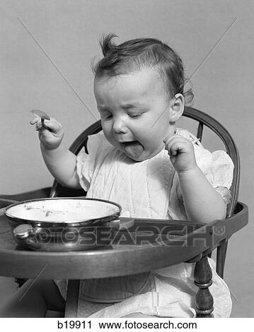 Stock Image - 1940S Baby Girl Eating Food In Highchair Sticking Out Tongue. Fotosearch