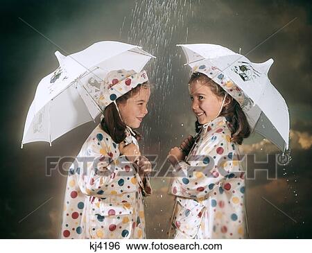 Stock Photograph - 1960S Smiling Twin Girls Wearing Matching Polka Dot Rain Coats And Hats Standing Under Umbrellas In The Rain. Fotosearch