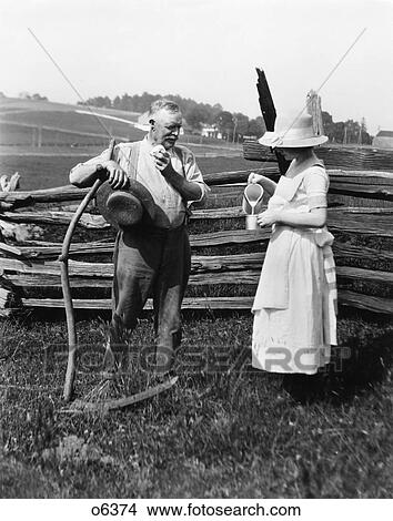Picture - 1890s 1900s 1910s WOMAN STRAW HAT APRON POURING GLASS MILK WATER FOR FARMER STANDING SWEATING BY FENCE LEANING ON SCYTHE. Fotosearch