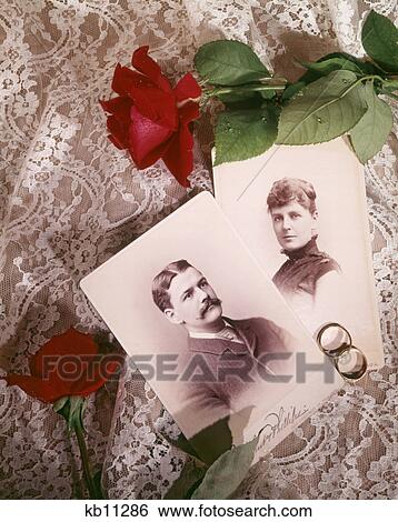 Stock Photograph - 1890s 1900s STILL LIFE OF ANTIQUE PHOTOGRAPHS HUSBAND WIFE COUPLE WITH WEDDING RINGS RED ROSES ON WHITE LACE. Fotosearch