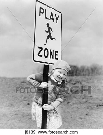 Stock Photo - 1920s 1930s SMILING GIRL LOOKING AT CAMERA HOLDING ON TO PLAY ZONE STREET SIGN. Fotosearch