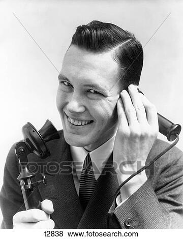 Stock Photo - 1920s 1930s SMILING MAN TALKING INTO CANDLESTICK TELEPHONE LOOKING AT CAMERA. Fotosearch