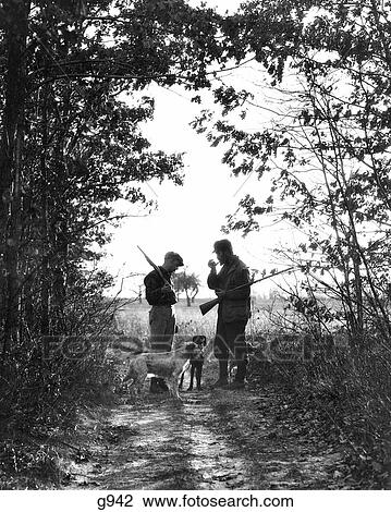 Stock Image - 1920s 1930s TWO MEN HUNTERS ANONYMOUS SILHOUETTED WITH DOGS ON COUNTRY LANE. Fotosearch