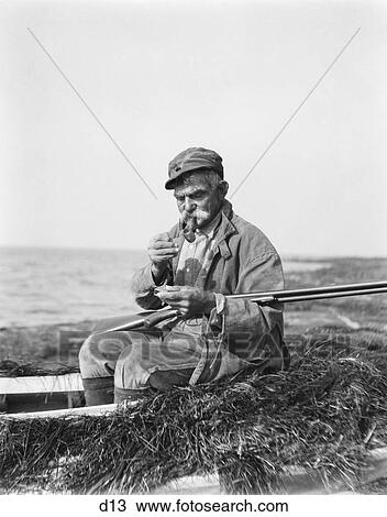1920S Man Lighting Corn Cob Pipe Sitting In Barnegat Bay Sneakbox Boat In Marshes Holding Shotgun Preparing To Go Duck Hunting View Large Photo Image Stock Image - 1920S Man Lighting Corn Cob Pipe Sitting In Barnegat Bay Sneakbox Boat In Marshes Holding Shotgun Preparing To Go Duck Hunting. Fotosearch