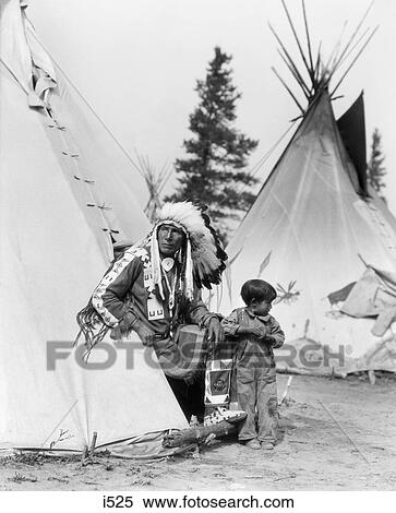 Stock Photography - 1920s NATIVE AMERICAN INDIAN MAN CHIEF SITTING EAGLE AND BOY IN ENTRANCE TO TEPEE STONEY TRIBE OF BANFF ALBERTA CANADA. Fotosearch