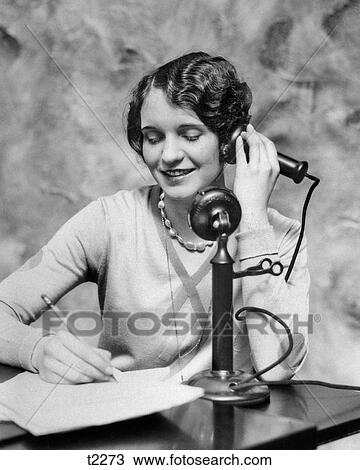 Stock Image - 1920s SMILING WOMAN SITTING AT DESK WRITING AND TALKING ON CANDLESTICK TELEPHONE. Fotosearch