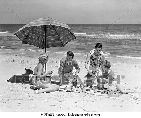 Stock Photo - 1930 1930s TWO COUPLES TWO MEN TWO WOMEN HAVING PICNIC ON BEACH BY OCEAN WITH TERRIER DOG UNDER STRIPED UMBRELLA. Fotosearch