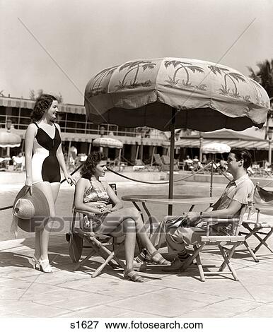 1930s 1940s 2 WOMEN BATHING SUITS SINGLE MAN CASUAL CLOTHES SITTING TALKING UNDER POOL SIDE UMBRELLA MIAMI BEACH FLORIDA USA View Large Photo Image Stock Photo - 1930s 1940s 2 WOMEN BATHING SUITS SINGLE MAN CASUAL CLOTHES SITTING TALKING UNDER POOL SIDE UMBRELLA MIAMI BEACH FLORIDA USA. Fotosearch