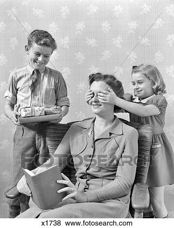 Stock Photo - 1930s 1940s LITTLE GIRL HAND OVER MOTHER EYES BOY WITH WRAPPED PRESENTS SURPISE CHRISTMAS OR MOTHER'S DAY. Fotosearch