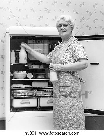 Stock Image - 1930s 1940s SENIOR WOMAN HOUSEWIFE GRANDMOTHER LOOKING AT CAMERA TAKING TWO GLASS BOTTLES OF MILK OUT OF KITCHEN REFRIGERATOR. Fotosearch