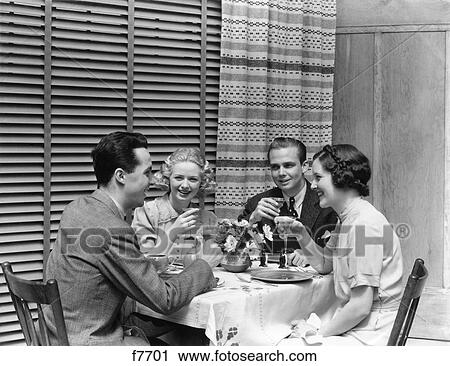 Stock Image - 1930s 1940s TWO COUPLES MAN WOMAN AT DINING TABLE HAVING COCKTAILS TOASTING RESTAURANT VENETIAN BLINDS CURTAINS. Fotosearch