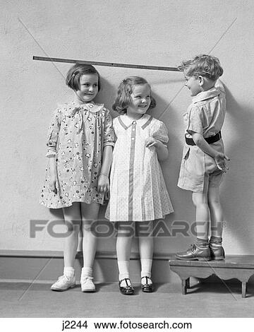 1930s BOY STANDING ON A STOOL MEASURING THE HEIGHT OF TWO GIRLS WITH YARD STICK View Large Photo Image Picture - 1930s BOY STANDING ON A STOOL MEASURING THE HEIGHT OF TWO GIRLS WITH YARD STICK. Fotosearch