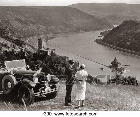 Stock Image - 1930s COUPLE WITH TOURING CAR OVERLOOKING RHINE RIVER BURG GUTENFELS AND CASTLE PFALZGRAFENSTEIN ON FALKENAU ISLAND GERMANY. Fotosearch