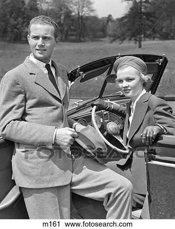 Stock Image - 1930s COUPLE WOMAN SITTING IN DRIVERS SEAT OF CONVERTIBLE CAR MAN LEANING ON OPEN DOOR HOLDING HAT. Fotosearch