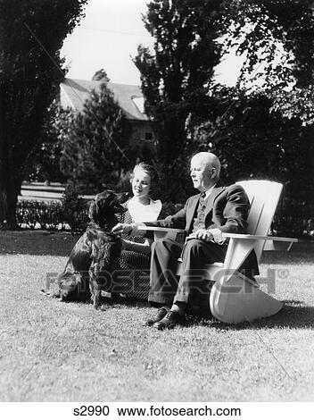 Stock Image - 1930s ELDERLY MAN SITTING ON ADIRONDACK CHAIR IN YARD YOUNG WOMAN KNEELING NEXT TO HIM WITH IRISH SETTER DOG. Fotosearch
