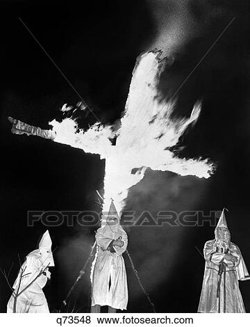Stock Photo - 1930s HOODED MEMBERS OF KKK KU KLUX KLAN STANDING IN FRONT OF BURNING CROSS. Fotosearch