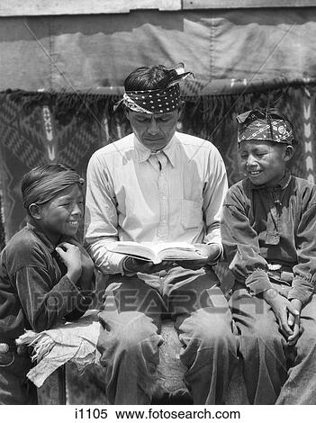 Stock Photography - 1930s NATIVE AMERICAN INDIAN NAVAJO MAN FATHER READING BOOK TO TWO SMILING BOYS SONS. Fotosearch