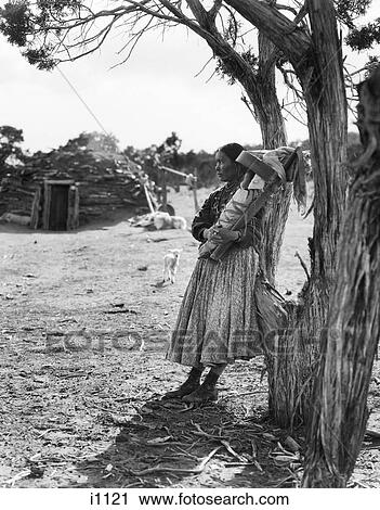 Stock Image - 1930S Native American Navajo Woman Holding Baby Papoose Under Trees With Hogan In Background. Fotosearch