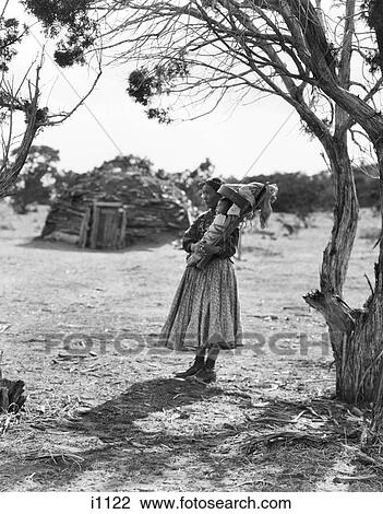 Stock Image - 1930S Native American Navajo Woman Holding Baby Papoose Under Trees With Hogan In Background. Fotosearch