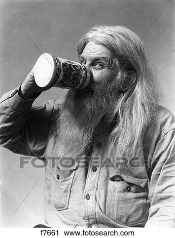 Stock Image - 1930s SENIOR MAN CHARACTER WITH SMILING EYES LOOKING AT CAMERA HIRSUTE LONG GRAY BEARD AND HAIR DRINKING BEER FROM STEIN. Fotosearch