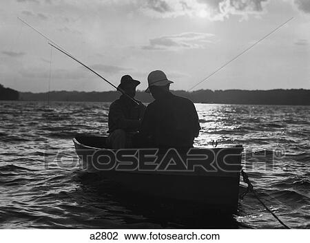 Stock Image - 1930S Two Anonymous Men Wearing Hats In Rowboat Fishing Silhouetted At Sunrise. Fotosearch