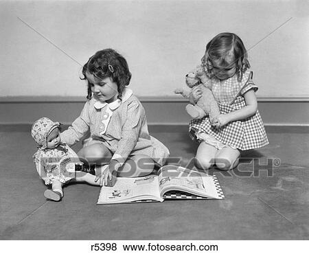 Stock Photo - 1930S Two Girls Sitting On Floor Playing One Girl Reading Book To Doll Other Holding Teddy Bear. Fotosearch