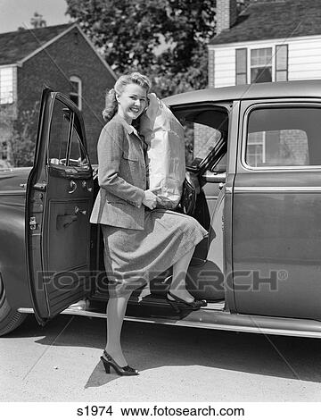 Picture - 1940s 1950s SMILING BLONDE WOMAN LOOKING AT CAMERA GETTING iNTO CAR HOLDING LARGE GROCERY BAG. Fotosearch