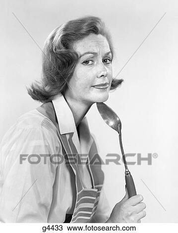 Stock Image - 1950s 1960s HOUSEWIFE RESTING CHIN ON MIXING KITCHEN SPOON WITH EXASPERATED TIRED FACIAL EXPRESSION LOOKING AT CAMERA. Fotosearch