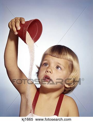 Stock Photography - 1950s 1960s LITTLE BLOND GIRL WEARING BATHING SUIT POURING SAND OUT OF RED PAIL. Fotosearch