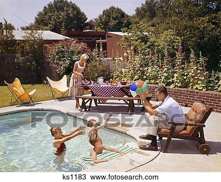 Stock Image - 1950S 1960S Man Father Tossing Ball To Three Children In Swimming Pool Mother Fixing Picnic Lunch Suburban Backyard Patio. Fotosearch