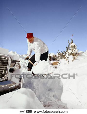 Stock Photograph - 1950s 1960s MAN WEARING RED CAP DIGGING OUT HIS CAR SHOVELING SNOW IN DRIVEWAY. Fotosearch