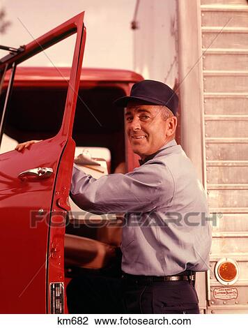 Stock Image - 1950s 1960s SMILING MAN DELIVERY DRIVER WEARING CAP AND UNIFORM LOOKING AT CAMERA STEPPING INTO CAB THROUGH OPEN TRUCK DOOR. Fotosearch