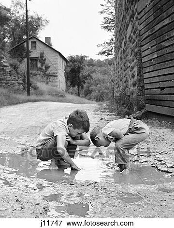 1950s 1960s TWO BOYS BENDING LOOKING AT PLAYING IN MUD PUDDLE View Large Photo Image Stock Photo - 1950s 1960s TWO BOYS BENDING LOOKING AT PLAYING IN MUD PUDDLE. Fotosearch