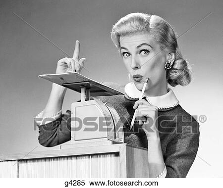 Stock Photography - 1950s BLOND WOMAN SECRETARY WEIGHING MAIL ON POSTAGE SCALE POINTING FINGER SURPRISED EXPRESSION. Fotosearch