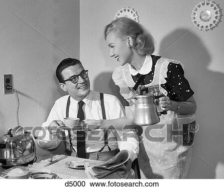 Stock Image - 1950s COUPLE AT BREAKFAST TABLE MAN READING NEWSPAPER SMILING AT WIFE HOLDING COFFEE CUP AND PERCOLATOR POT. Fotosearch