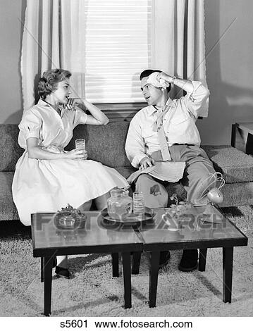 Stock Image - 1950s COUPLE SITTING ON LIVING ROOM COUCH TRYING TO COOL DOWN DRINKING ICED DRINK BEFORE FAN ON COFFEE TABLE. Fotosearch
